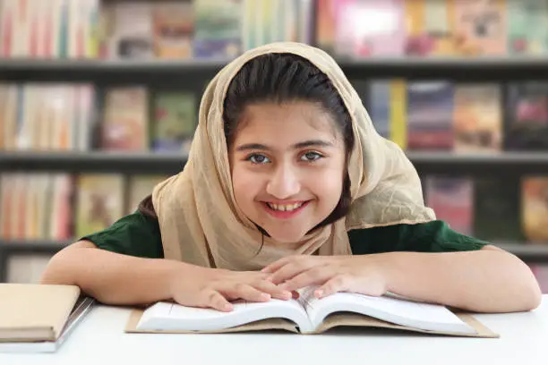 Adorable smiling Pakistani Muslim girl with beautiful eyes wearing hijab, studying and doing homework on table, happy student kid reading book on blurred background of bookcase in library.