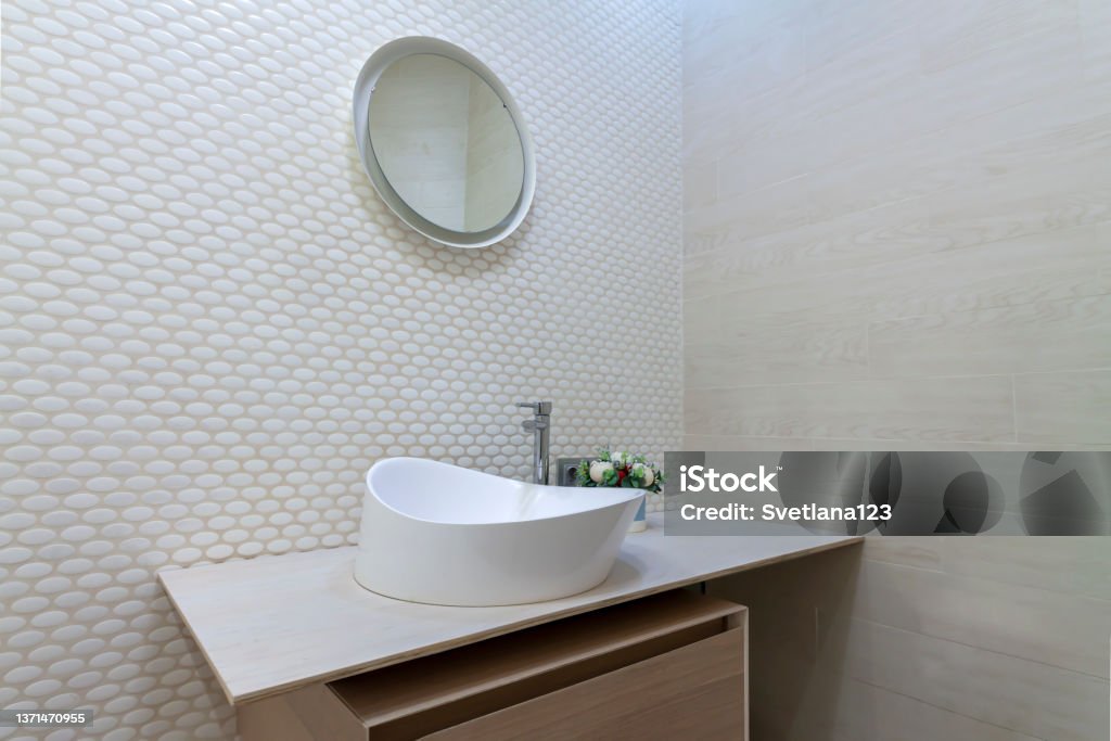 White washbasin with faucet on wooden countertop in minimalist modern bathroom. Scandinavian interior with tiled mosaic wall wall and round mirror. Copy space and nobody. White washbasin with faucet on wooden countertop in minimalist modern bathroom. Scandinavian interior with tiled mosaic wall wall and round mirror. Copy space and nobody in the house. Bathroom Sink Stock Photo