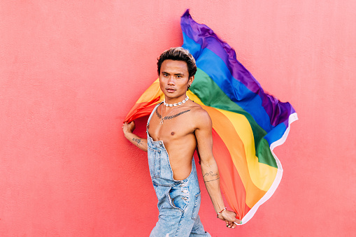 Portrait of a handsome young man holding pride movement LGBT rainbow flag against a pink wall. Man with a pride flag looking at camera outdoors.