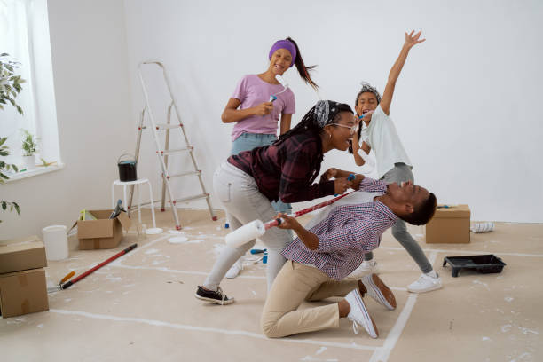 la familia organiza una fiesta durante la renovación del apartamento, tomando un descanso de pintar paredes, los padres con niños cantan a los rodillos, bailan, el hombre cae de rodillas y finge tocar la guitarra - home improvement couple painting real estate fotografías e imágenes de stock