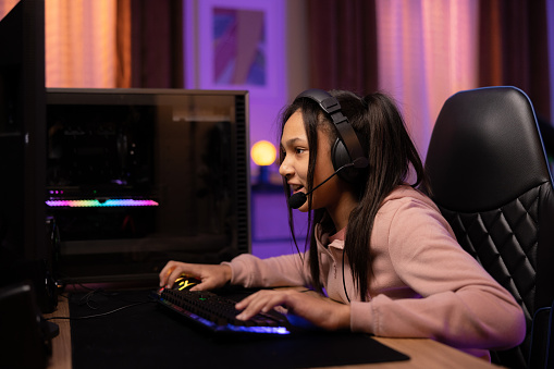 Joyful professional gamer girl wearing headphones with microphone playing online video games chatting with friends team members colorful neon led purple lights in room