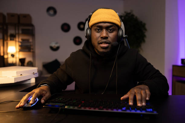 istock Seroious man in a yellow cap with a headset is playing a video shooter carefully on a computer. Online multiplayer championship. In the background a dark room at night with a mess of pizza boxes 1371486576