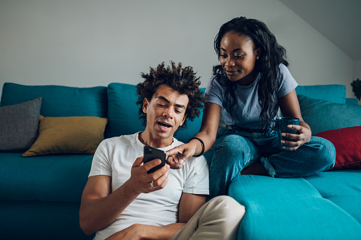 Cheerful african american couple spending day off at home together while using smartphone and drinking coffee. Romantic couple drinking coffee at home.