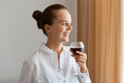 Side view portrait of smiling positive woman wearing white shirt holding glass of red wine, looking away with toothy smile, expressing happiness, celebrating festive event.