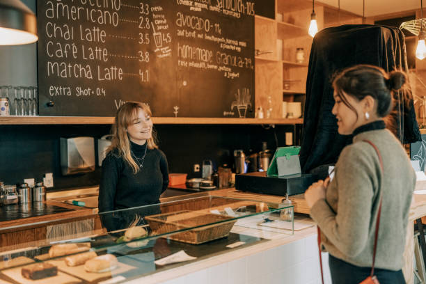 Smiling young woman barista talking with a female customer while standing behind the counter of a cafe. Waitress taking order from customer in coffee shop.