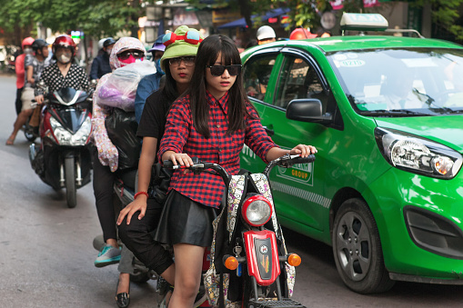Two girls on a motor scooter not wearing crash helmets, mix it with cars and other traffic on Bà Triệu, Tràng Tiền, Hoàn Kiếm, Hanoi, Vietnam