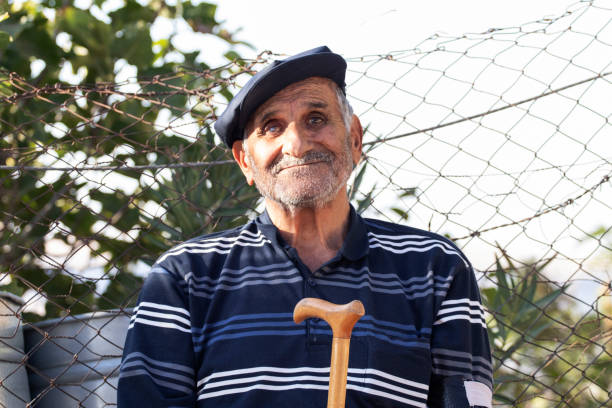 A close-up, wide-angle portrait of a 64-year-old man with white hair and a light beard, wearing a large blue hat, captured outdoors. He is striking playful and fun poses, with a mischievous smile, and the cheerful vibe of his oversized blue hat adds a whimsical touch. Natural light enhances the details of his face and the vibrant color of the hat. The blurred background gives the photo a fresh and energetic atmosphere.
