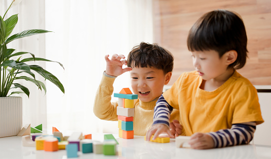 Two little Asian boys on the balcony of their home. They were wearing yellow sweaters and playing with blocks at the dining table. By the window, the bright and soft sunlight came in, and they had a great time.