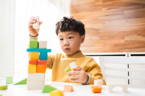 A little asian boy, on the balcony of his home. Wearing a yellow sweater and curly hair, he was playing with building blocks on the dining table. Next to the window, the bright and soft sunshine came in, and he had a good time.
