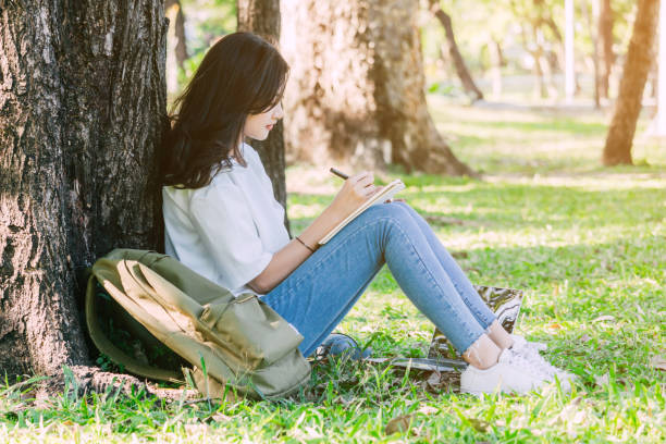 Woman with pen writing on a notebook sitting near the tree in park stock photo
