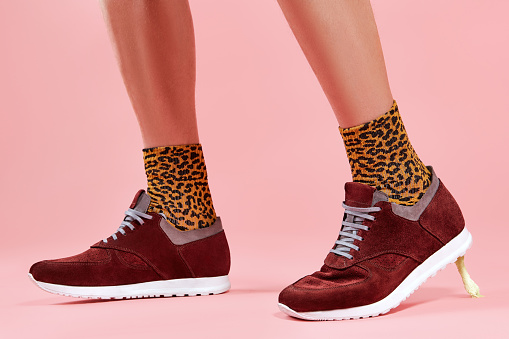 Closeup of male legs in leopard print socks and maroon suede sneakers with chewing gum sticking to white sole on pink studio background, cropped shot