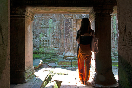Young woman silhouetted between massive pillars within a stone wall reveal of ornately carved temple walls, Ta Prohm, Angkor Wat, Cambodia, Southeast Asia, 28th September 2019. A female visitor in bright loose fitting clothes stands in quiet contemplation of the ornately carved walls of the inner temple of Ta Prohm  - one of many devotees who sit or wander around the ruins of the ancient Angkor Wat stone temple complex