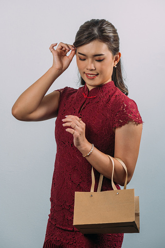 Happy young beautiful Asian woman wearing red oriental Cheongsam dress with luxury accessories such as golden ring, earring and bracelet holding shopping bag with white background.
