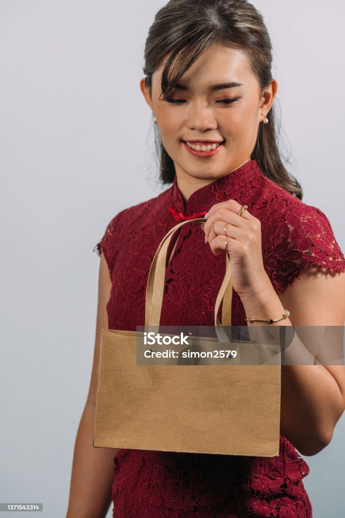Happy young beautiful Asian woman wearing red oriental Cheongsam dress with luxury accessories such as golden ring and bracelet with white background. Happy young beautiful Asian woman wearing red oriental Cheongsam dress with luxury accessories such as golden ring, earring and bracelet holding shopping bag with white background. 30-34 Years Stock Photo