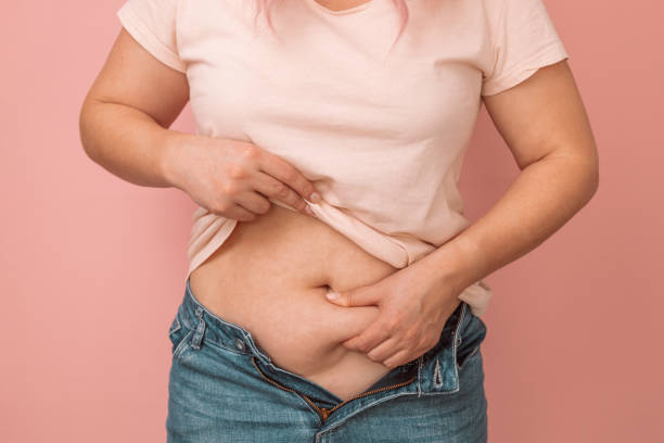Woman hand holding excessive belly fat, overweight concept. Overweight female wearing a jeans Close up of woman hand holding excessive belly fat isolated on pink background, overweight concept human abdomen stock pictures, royalty-free photos & images