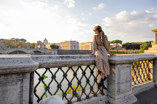 Woman enjoying beautiful view of Rome city, sitting on the bridge during the morning time. Elegant woman wearing dress and shawl in hair. Concept of italian lifestyle and travel