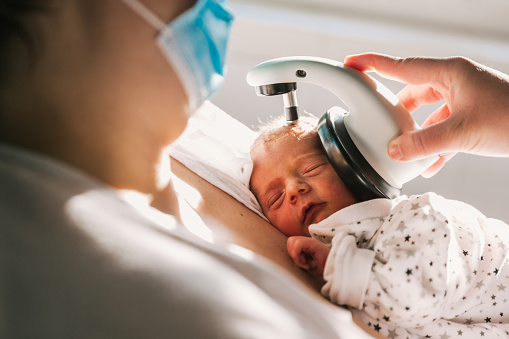 From above crop mom in face mask using modern device to check hearing of sleeping infant in sunlit ward of hospital