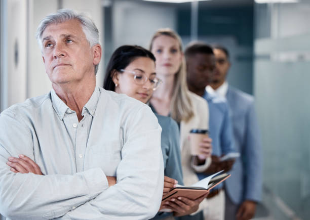 Shot of a mature businessman standing with his arms crossed while waiting in line in an office How much longer do I have to wait? impatient stock pictures, royalty-free photos & images