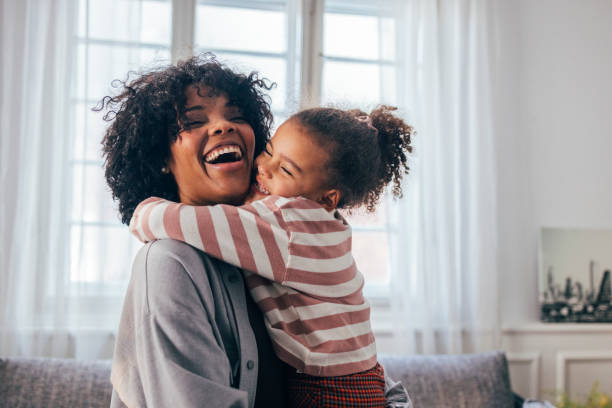 una feliz mujer afroamericana siendo abrazada por su linda hija pequeña en casa - madre fotografías e imágenes de stock