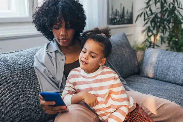 Photo of An Adorable African-American Child Sitting On The Sofa Watching A Cartoon On The Mobile Phone Her Mother Is Holding