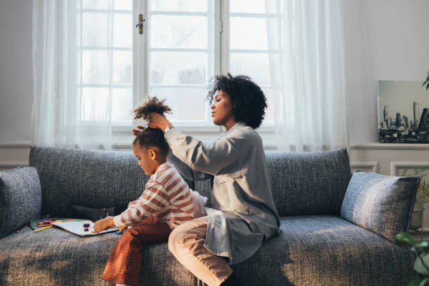 una vista laterale di una madre afro-americana che fa i capelli della sua graziosa bambina mentre sono seduti sul divano di casa - art indoors lifestyles education foto e immagini stock