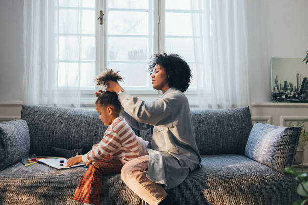 a side view of an african-american mother doing her cute little daughter's hair while they are sitting on the sofa at home - education relaxation women home interior imagens e fotografias de stock