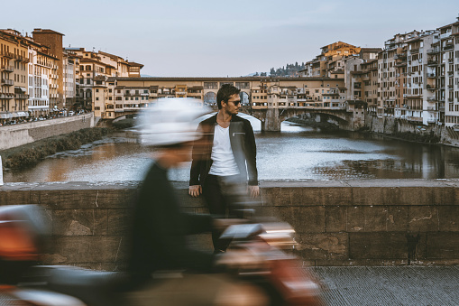 Portrait of a happy young handsome traveller man with sun glasses and sitting, smiling, having fun outdoor at the coastline of Arno river and Ponte Vecchio in the background at a sunset time in Florence Old City, Tuscany, Italia