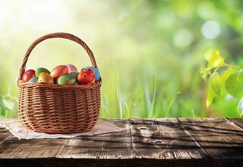 Wicker basket with Easter colorful eggs on rustic wooden table with defocused lush foliage at background. Backdrop for product display on top of the table. Spring Easter composition with copy space.
