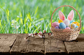 Easter eggs in basket and Easter bunny ears behind a basket on empty wooden table.