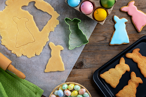 Process of making Easter gingerbread cookies in the shape of an Easter bunny from dough on a wooden table. Easter greeting card with cooking gingerbread cookies and colorful Easter eggs and candies. Flat lay.