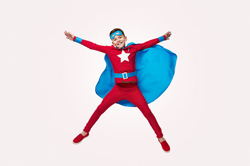 Delighted boy in superhero costume jumping above ground with outstretched arms on white background and looking at camera