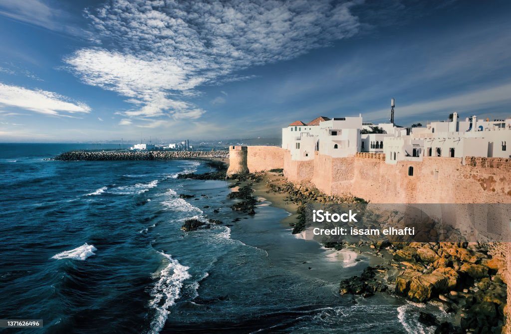 White Painted Historical Buildings And City Wall Of Asilah Against Blue Sky At Coast, Morocco, North Africa Asilah Stock Photo