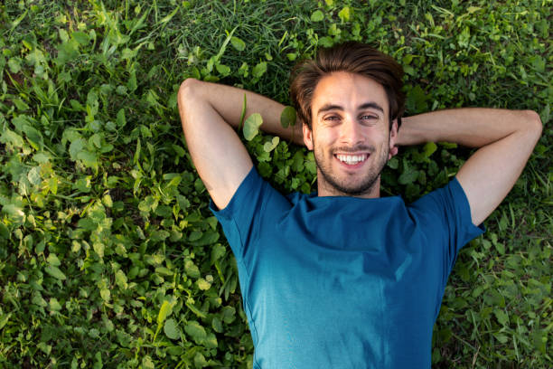 vue de dessus d’un jeune homme heureux et souriant se relaxant allongé sur l’herbe en regardant la caméra. espace de copie - hands behind head photos et images de collection