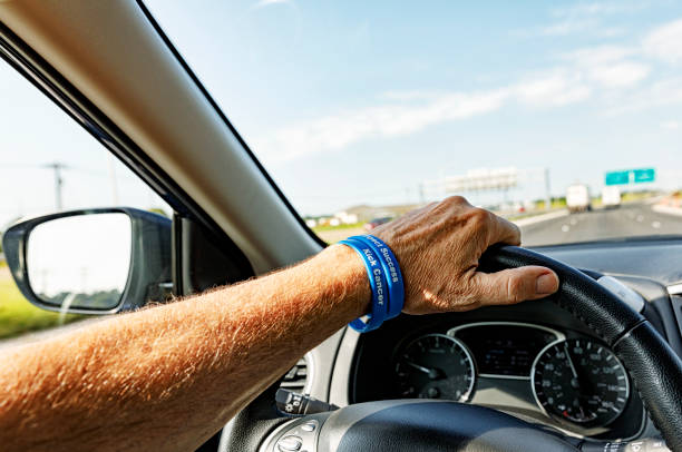 Senior man cancer survivor driver's hand on a car steering wheel. He is wearing two blue colon cancer support wristbands labeled: \