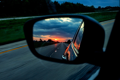 Reflection of the distant dawn morning sun rising into a dark cloudscape reflected in the rear view mirror of a car speeding westward on an expressway multiple lane highway just after sunrise.