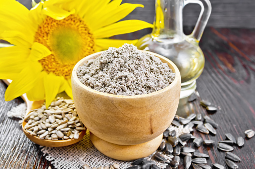 Sunflower flour in bowl, oil in decanter, seeds in a spoon on burlap, sunflower flower on wooden board background