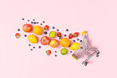 Shopping trolley with fruits on pink background, table top view