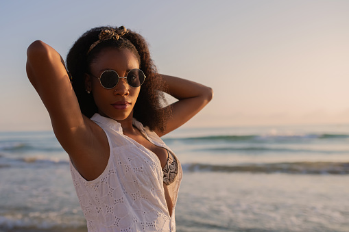 Woman wearing sunglasses on the beach in white summer clothes and bikini, looking at the camera with both hands behind head at sunset. Selective focus.