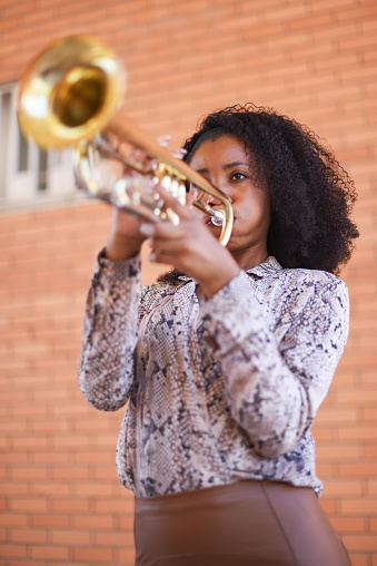 Vertical image of an african american woman standing with curly hair playing the trumpet with a brick wall background outside. Selective focus.