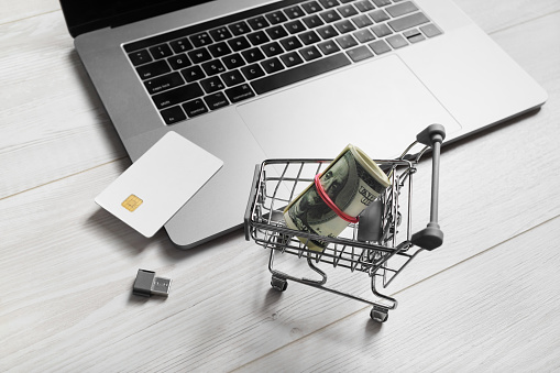 Online shopping concept. Laptop, shopping trolley, credit card, money and usb flash drive on light wooden background