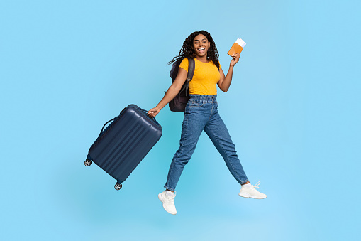 Emotional african american millennial woman in casual outfit with suitcase jumping up, holding flight ticket and passport, blue studio background, full length shot, panorama with copy space