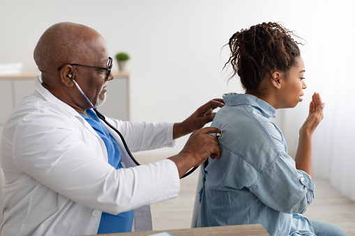 Pneumology Consultation Concept. Black male general practitioner examining coughing female patient in modern clinic, using stethoscope, listening lungs from the back. Medicine And Healthcare