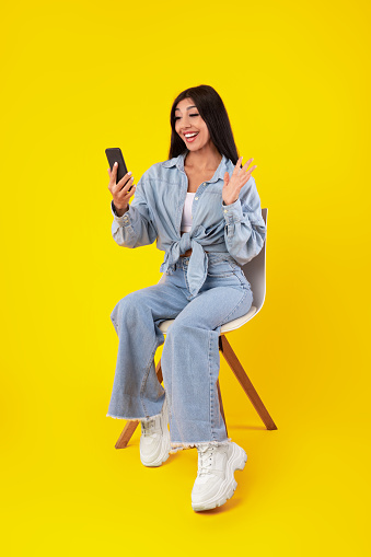 Video Conference. Cheerful young woman having web call using cell phone, talking waving hand at webcam, excited lady sitting on chair isolated over yellow studio background, vertical banner