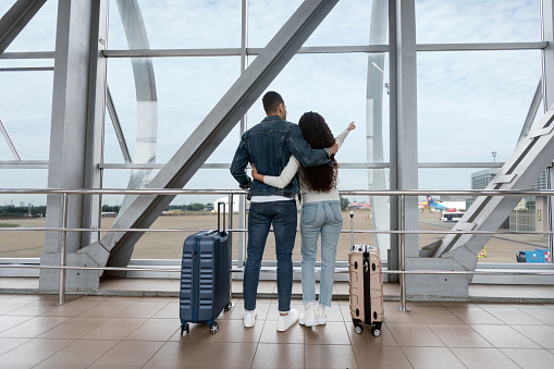 Air Journey. Romantic Couple Looking At Window In Airport And Pointing Away, Rear View Shot Of Young Spouses Standing With Suitcases At Terminal, Waiting For Flight, Ready For Vacation Trip