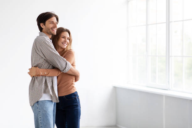 Cheerful millennial caucasian male and female hugging, looking at window in empty room of new apartment, sun flare stock photo