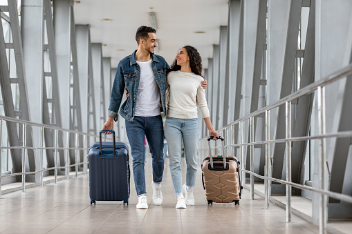Trip Together. Happy Young Arab Spouses Walking With Luggage At Airport Terminal, Romantic Middle Eastern Couple Embracing And Smiling To Each Other While Going To Boarding Gate, Copy Space