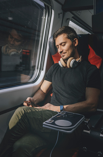 Happy young handsome man with BlueTooth headphones is travelling first class on a speedy train sitting, smiling, having fun, texting and reading messages while using his smartphone and looking out through the window