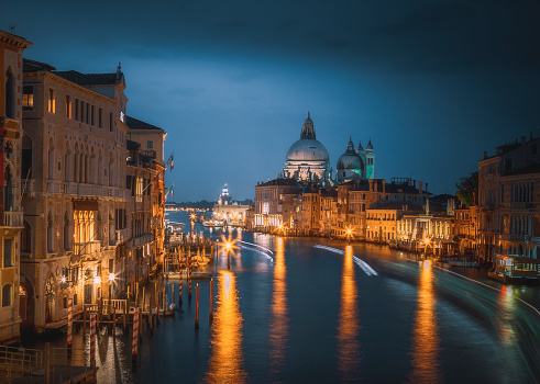 Beautiful illuminated landscape city view of famous Canal Grande with Basilica di Santa Maria Della Salute church. Old boats, gondolas and canoes moving on the river on a sunny day with blue sky in summer at night, Venice, Veneto, Italia.