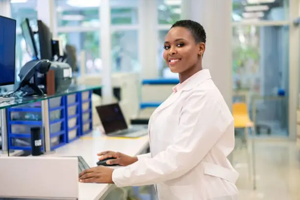 Portrait of a woman scientist working on a computer while standing at the table in a science lab and looking at camera. Female researcher wearing a lab coat working in a laboratory.