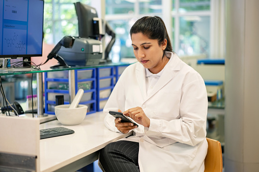 Asian woman scientist sitting at desk using her mobile phone. Female medical researcher wearing lab coat working at the laboratory.